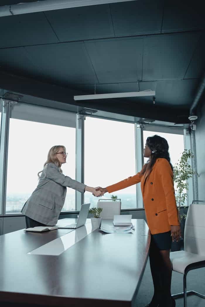 embark Two businesswomen shake hands across a conference table in a modern office setting.