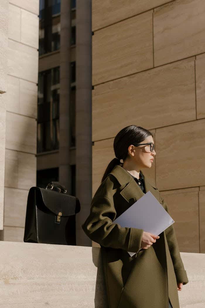 contact-bg Businesswoman in a coat holding documents outside an office building with a briefcase nearby.